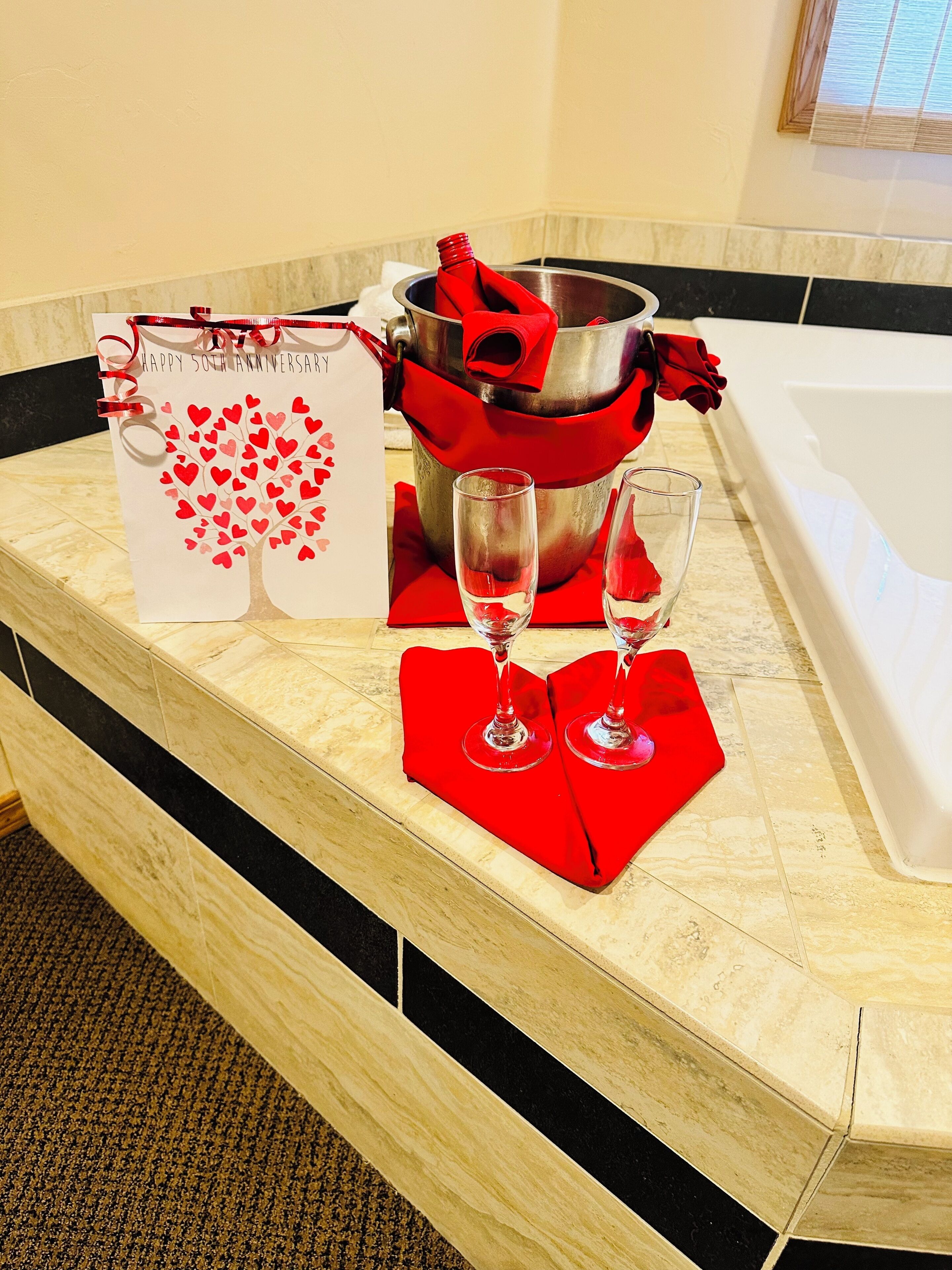 Habitación de hotel con encimera de mármol, una caja de regalo, un cubo con tela roja y dos copas de vino.