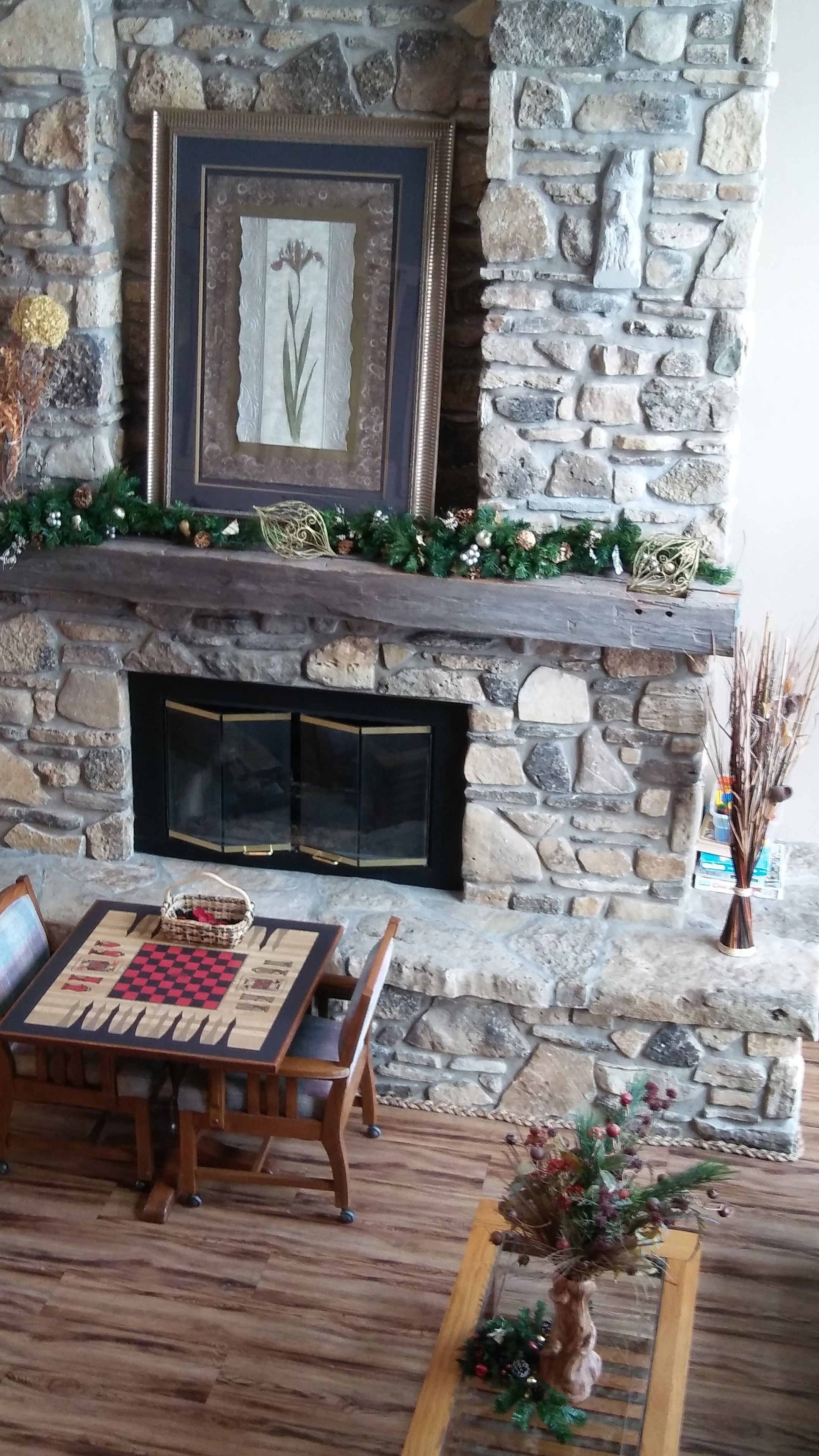 A stone fireplace with a framed artwork above it, a checkered table with chairs, and a decorative plant on the floor.