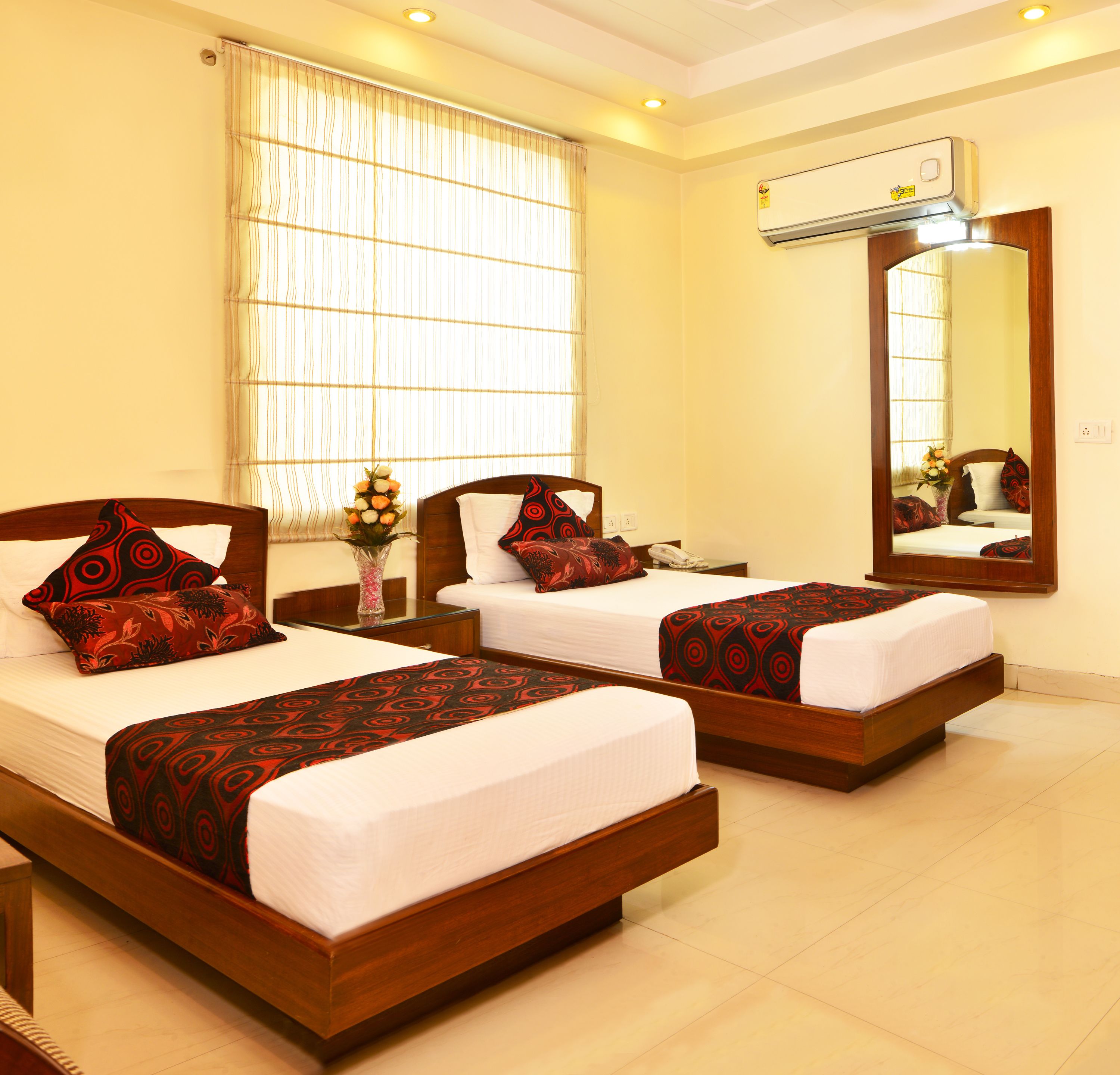 A hotel room with two beds, a large window with blinds, an air conditioning unit, and a mirror on the wall.