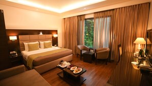 A hotel room with a large bed, a sitting area with a table, and a view of greenery outside.