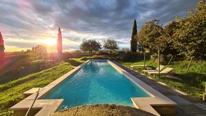 A rectangular pool with a wooden deck and lounge chairs, surrounded by greenery and trees, with a sunset in the background. - Maso di Villa (Susegana)