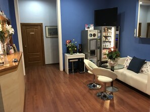 A room with a wooden floor, a white sofa, a small round table, a white chair, a vending machine, and a television on the wall. - Hotel Pelayo (Barcelona)