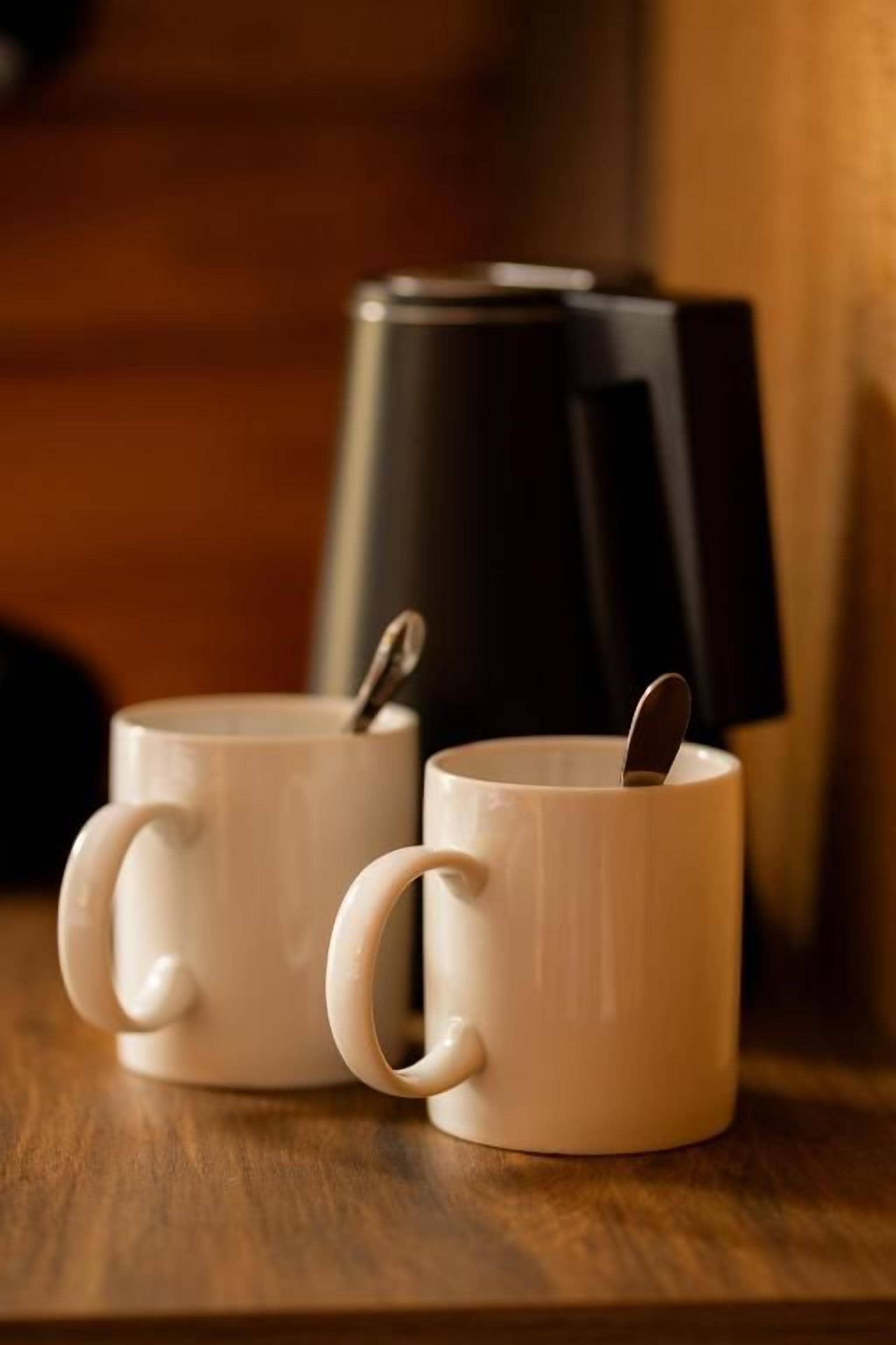 Dos tazas de cerámica blanca con asa, una de ellas contiene una cuchara. Están sobre una superficie de madera con un objeto negro al fondo.