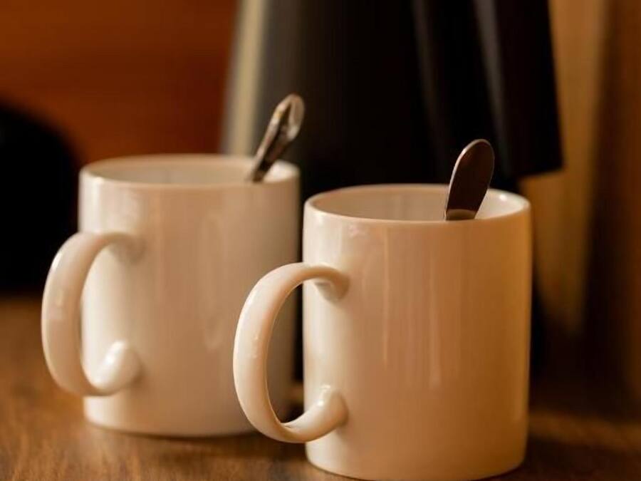 Two white ceramic mugs with handles, one of which contains a spoon. They are placed on a wooden surface with a black object in the background.