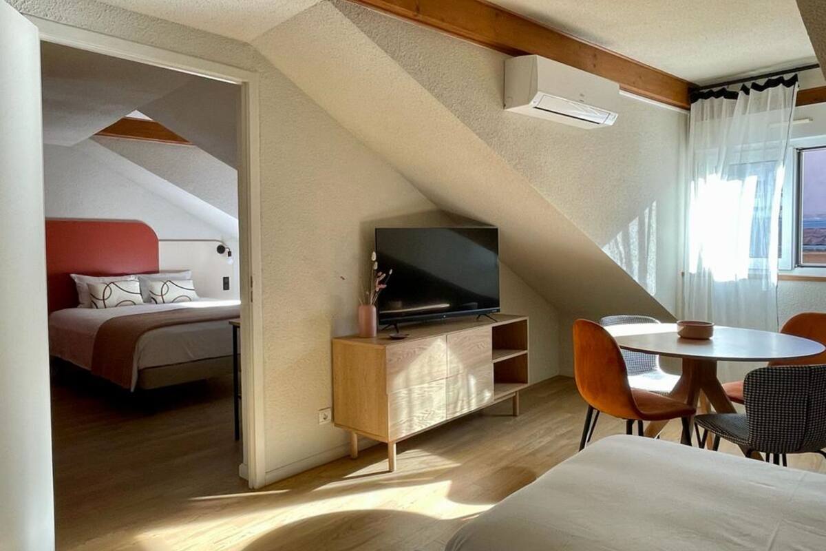 A compact room with a bed, a TV on a wooden cabinet, a dining table with chairs, and a window with curtains.
