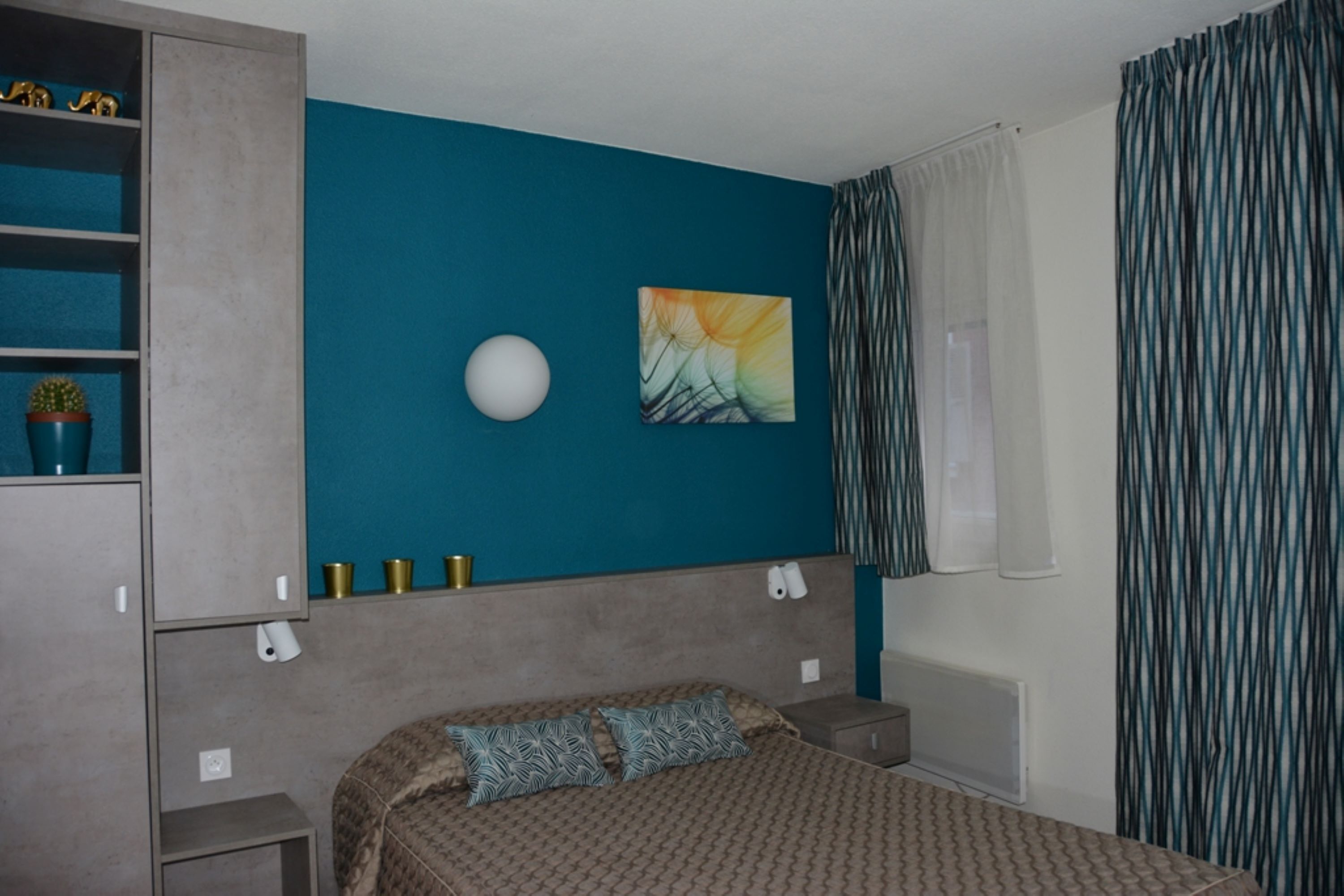 Une chambre avec un mur d’accent de couleur turquoise, un lit avec une literie à motifs, une table de chevet, une commode, une lampe fixée au mur et une fenêtre avec des rideaux.