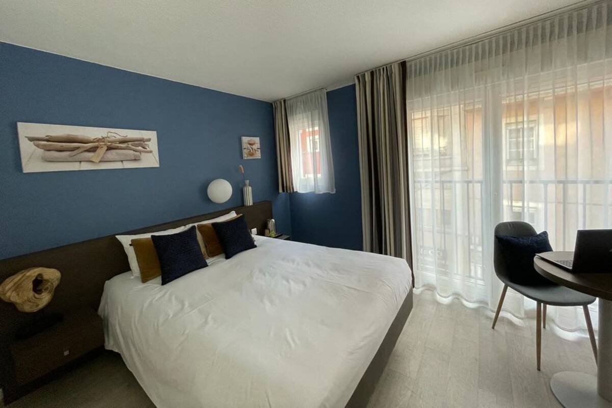 A hotel room with a large bed, a desk, and a chair. There is a window with curtains and a painting on the wall.