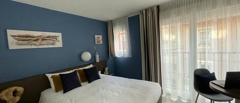 A hotel room with a large bed, a desk, and a chair. There is a window with curtains and a painting on the wall.