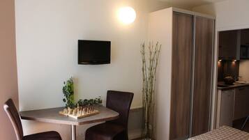 A compact living space with a dining table, two chairs, a television mounted on the wall, and a vertical plant beside a wardrobe.