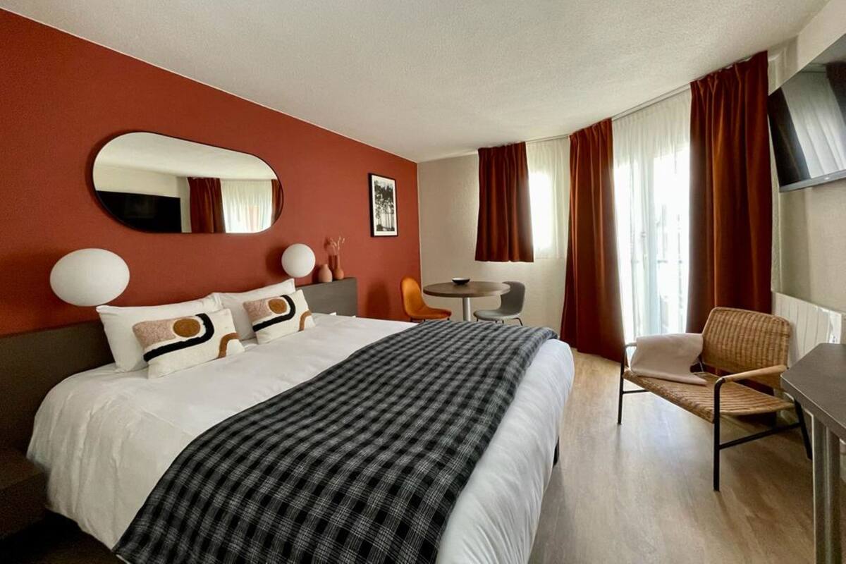 A hotel room with a large bed, a round table, a chair, a desk, a mirror, and a window with curtains.