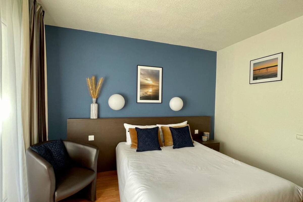 A hotel room with a bed, a chair, a nightstand, and a wall with a painting and two spherical wall lights.