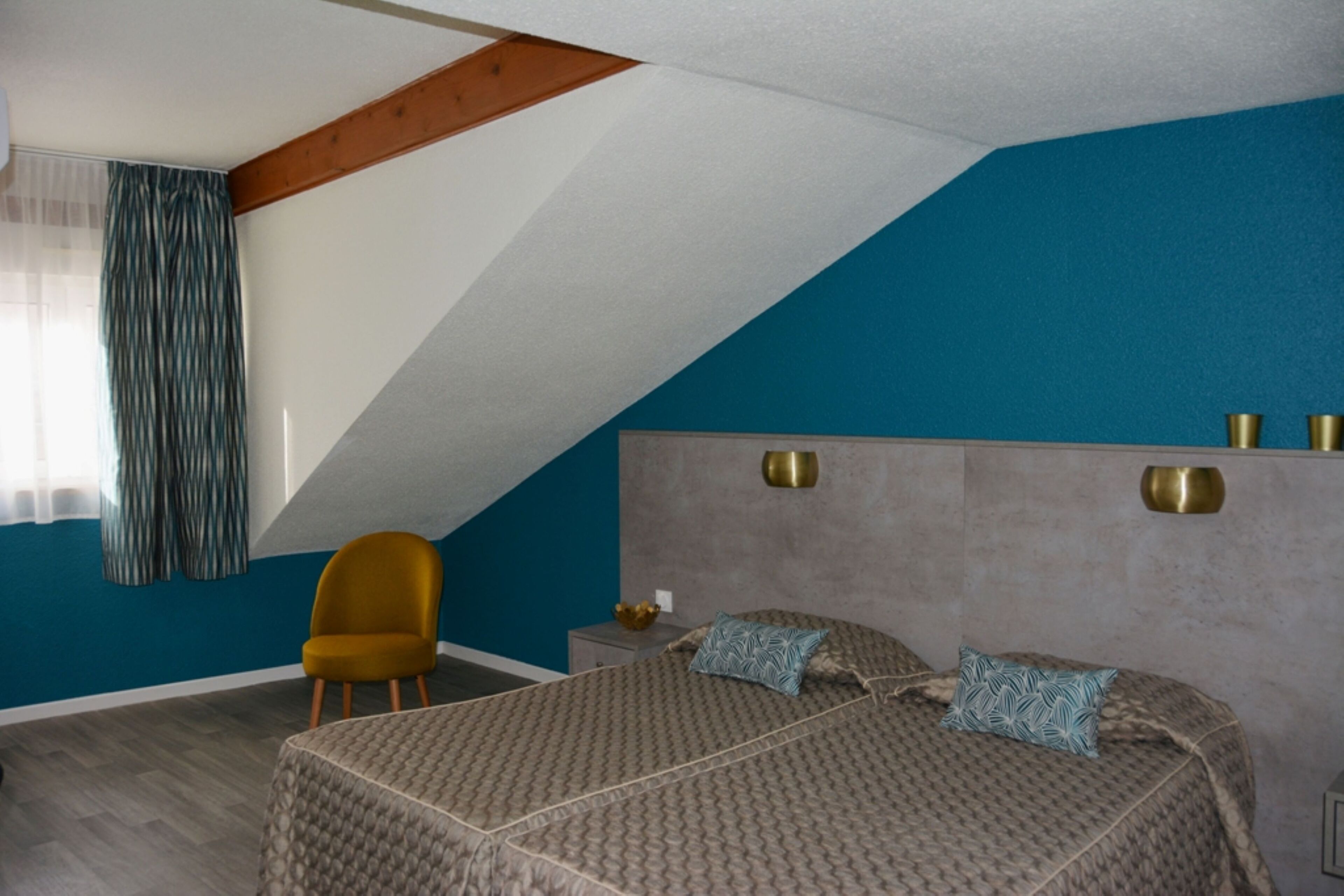Una camera da letto con soffitto spiovente, un letto con biancheria a motivi, una poltrona color senape e un comodino con una ciotola.