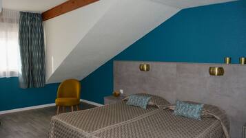 Una camera da letto con soffitto spiovente, un letto con biancheria a motivi, una poltrona color senape e un comodino con una ciotola.