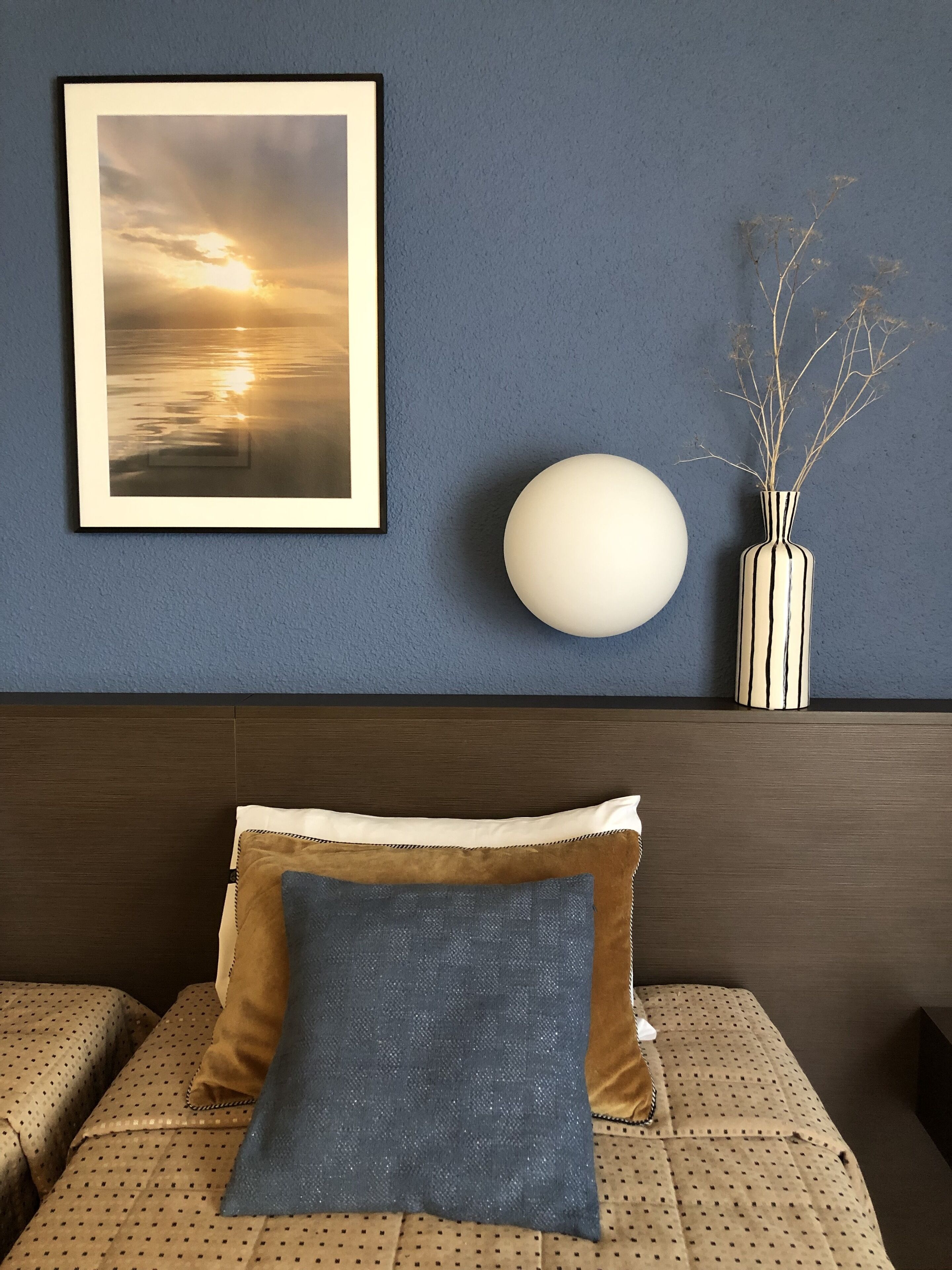 A room with a framed sunset photo, a white spherical light, and a vase with branches on a blue wall.