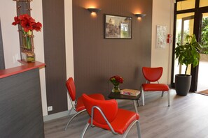 A reception area with a red counter, two red chairs, a small table with a vase of red flowers, and a framed picture on the wall. - Les Baladines Residence Tourisme (Thonon-les-Bains)
