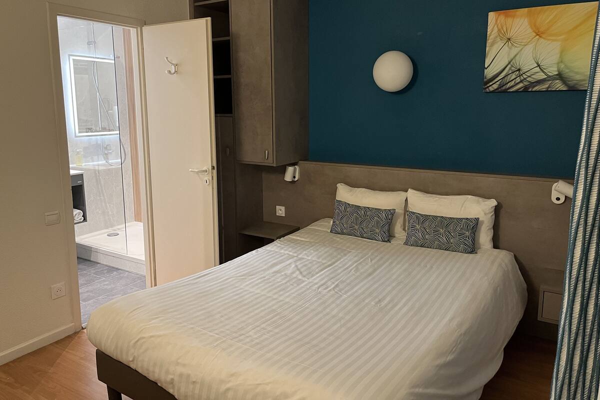 A bedroom with a bed, a wardrobe, a bathroom with a shower, and a wall-mounted light fixture.