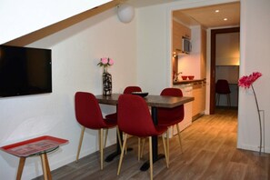 A dining area with a table and red chairs, a TV mounted on the wall, and a kitchen in the background. - Les Baladines Residence Tourisme (Thonon-les-Bains)