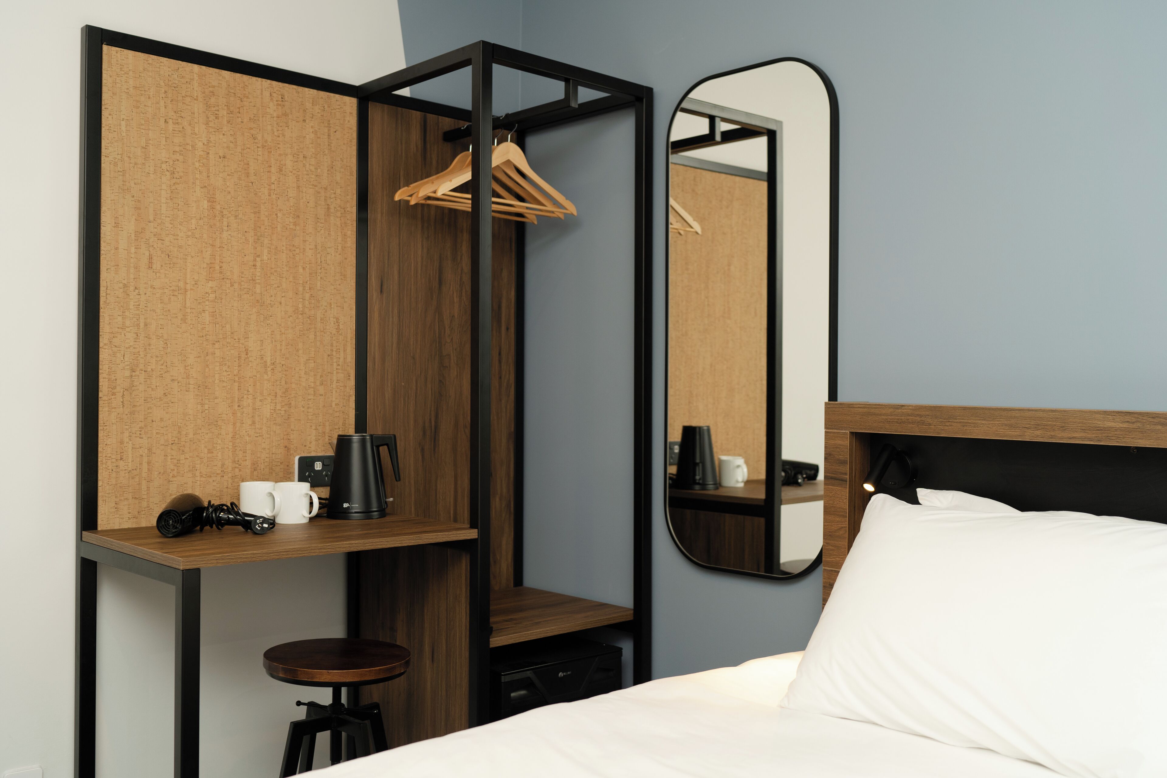 A modern hotel room with a wooden desk, a mirror, a bed, and a hanging coat rack.