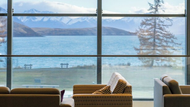 Una habitación moderna con ventanas amplias que ofrecen vistas a un lago y montañas. Hay dos sofás y una silla de mimbre.