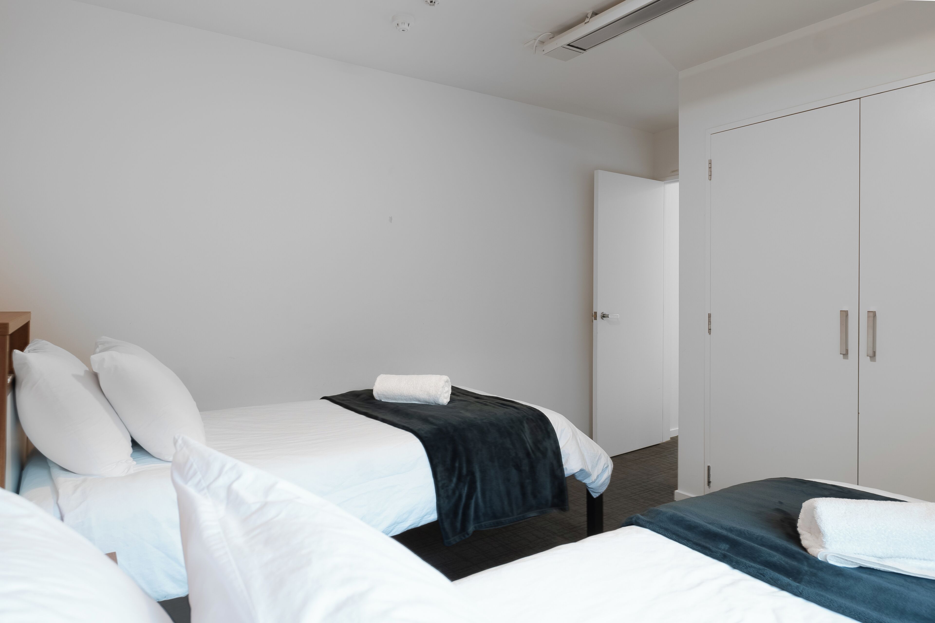A hotel room with two beds, white bedding, a dark blue blanket on one bed, and a white towel on the other. There is a wardrobe with two doors and a small mirror.
