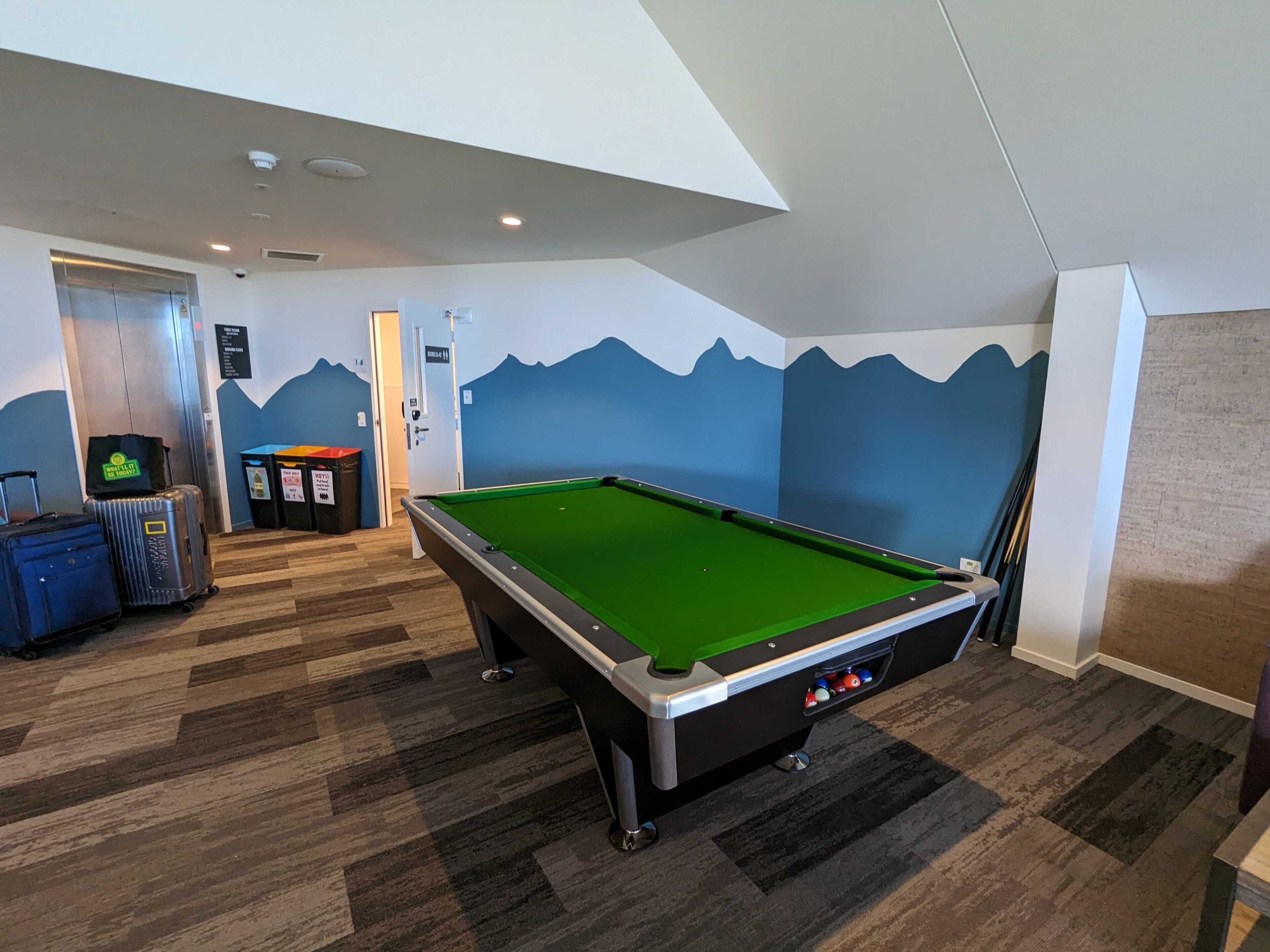 Une table de billard dans une pièce dont un mur est décoré d’une fresque représentant une montagne, avec des bagages et une poubelle.