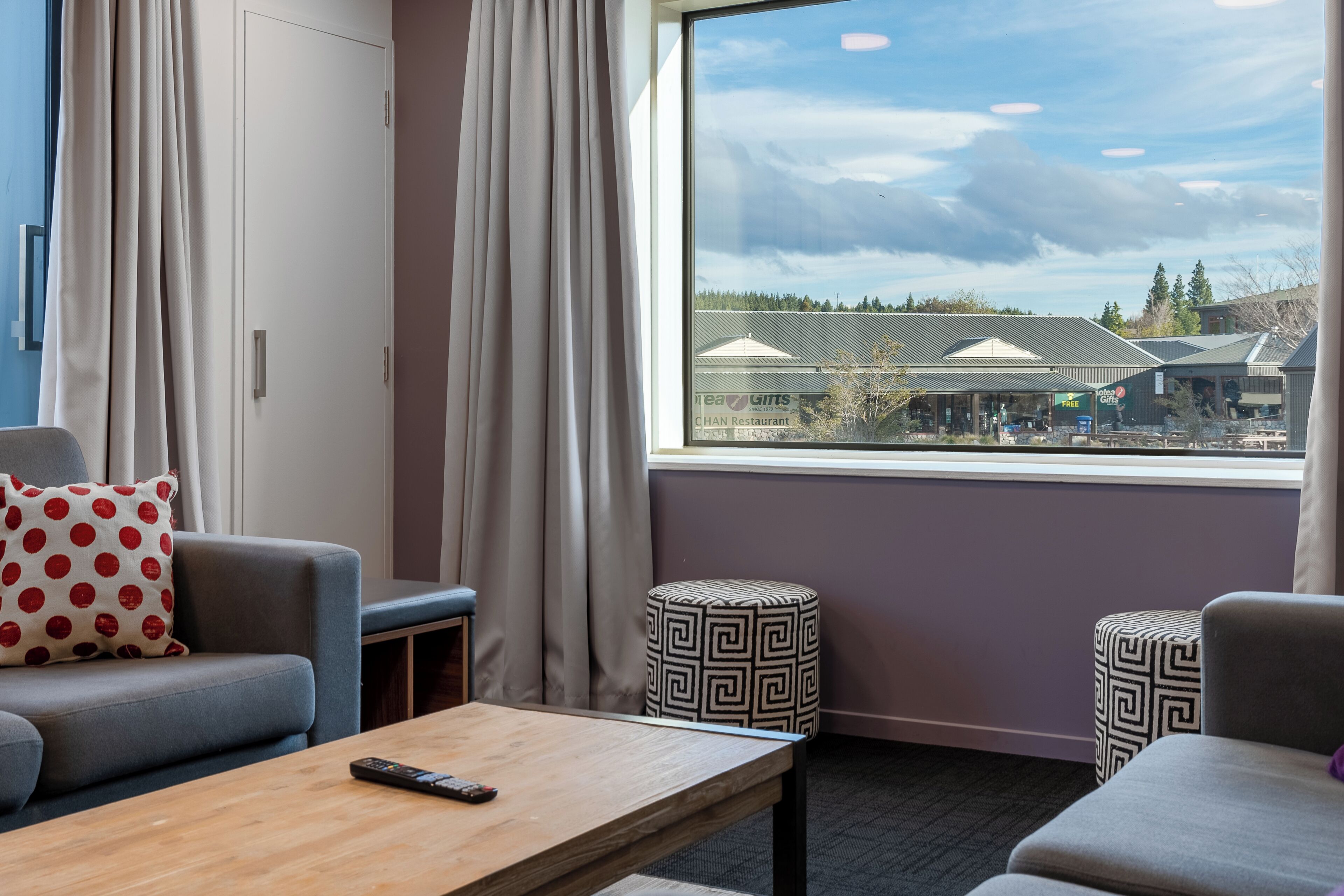 A hotel room with a large window, a sofa, a coffee table, and a patterned ottoman.