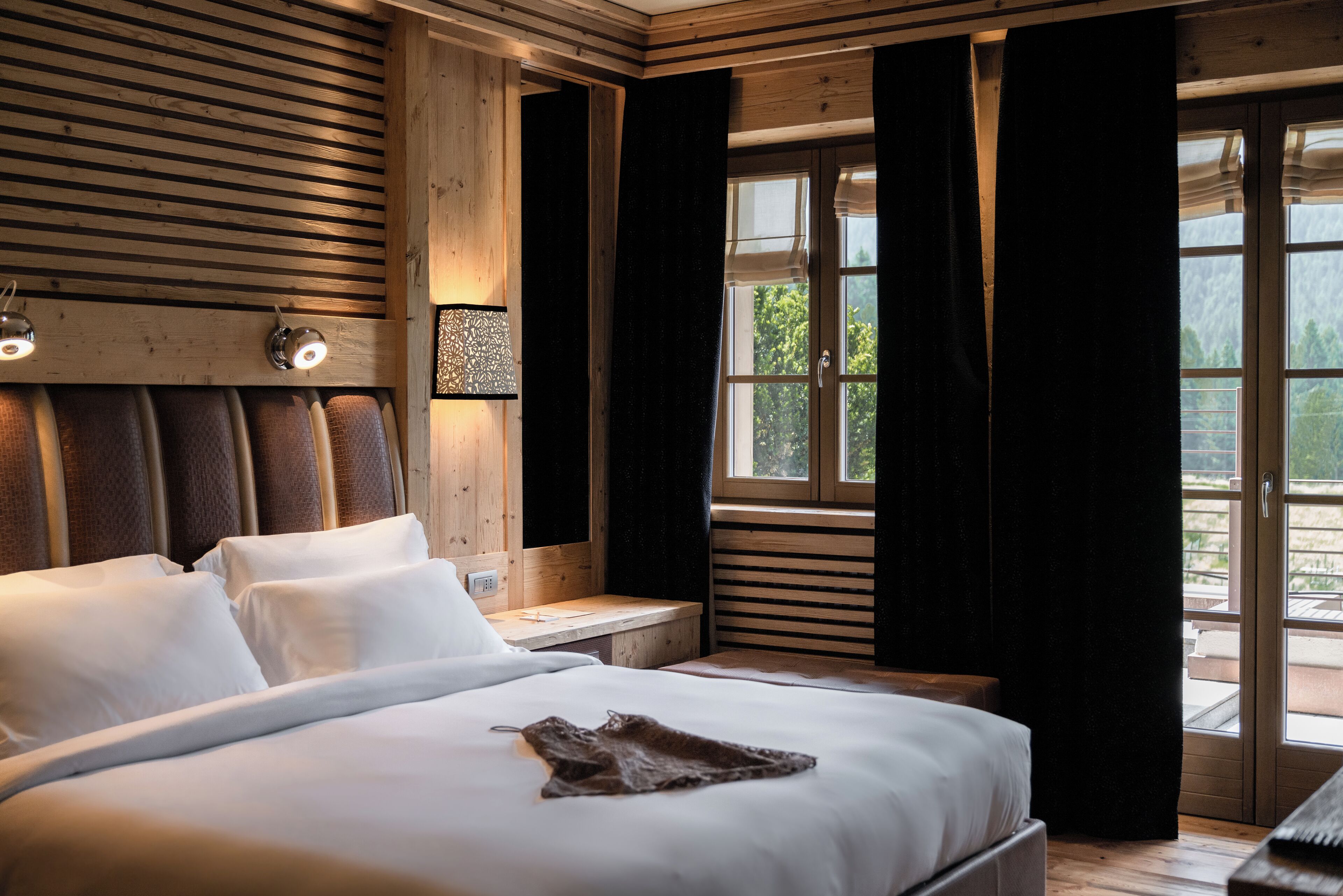 A bedroom with a wooden headboard, a large bed with white linens, a bedside table with a lamp, and a window with curtains.