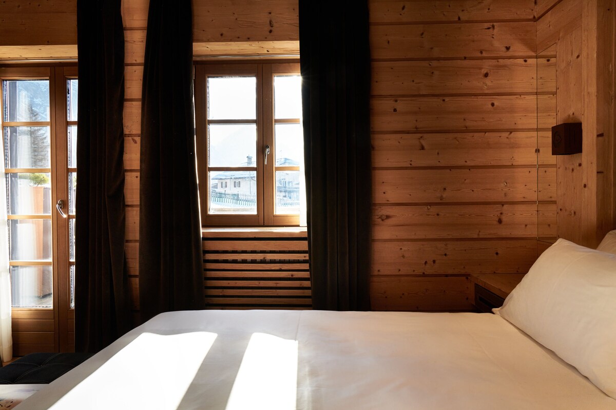 Habitación de cabaña de madera con cama, cortinas y ventana con vistas a edificios.