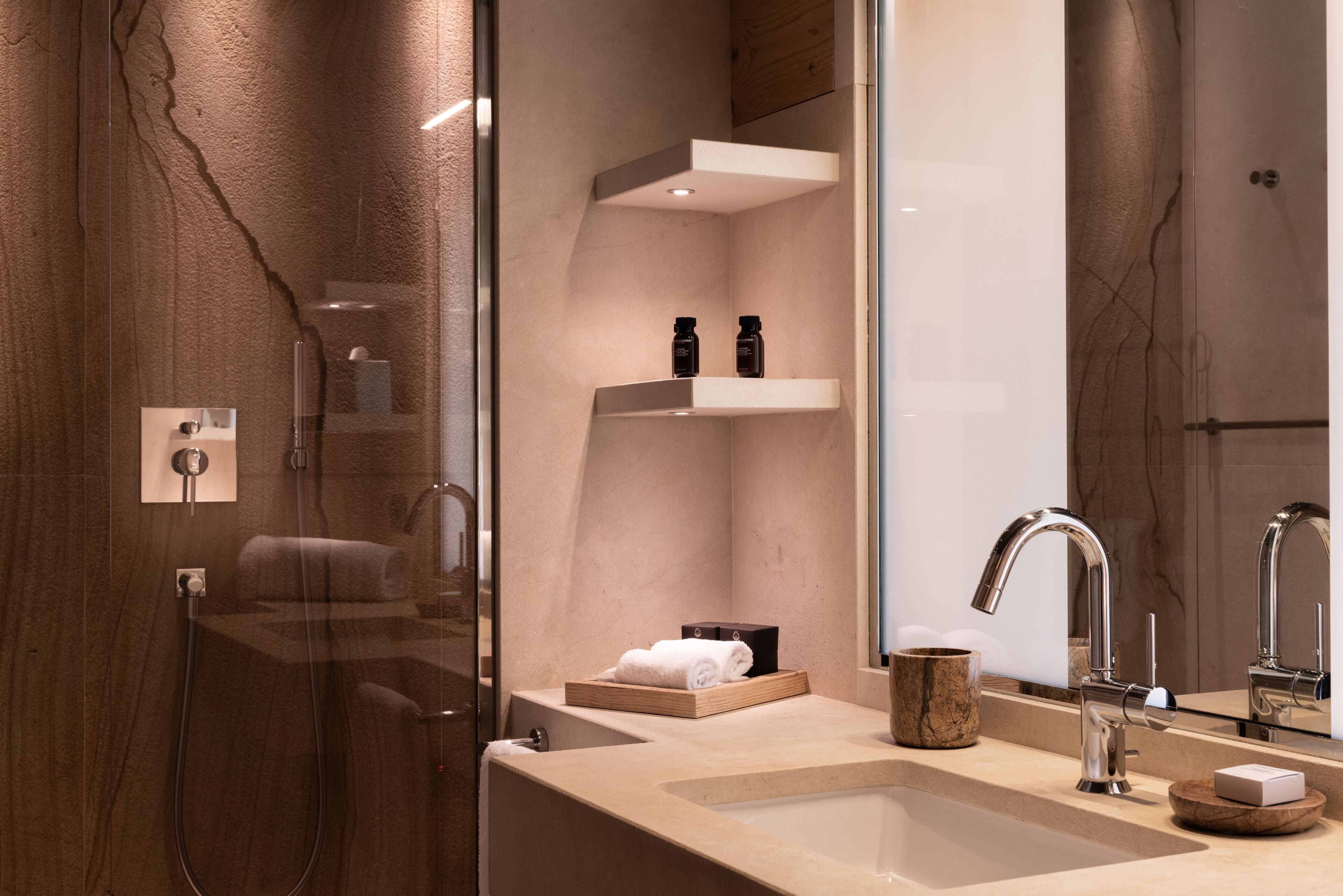 A modern bathroom with a large mirror, a sink with a faucet, and a shower area. There are shelves with toiletries and a towel rack.
