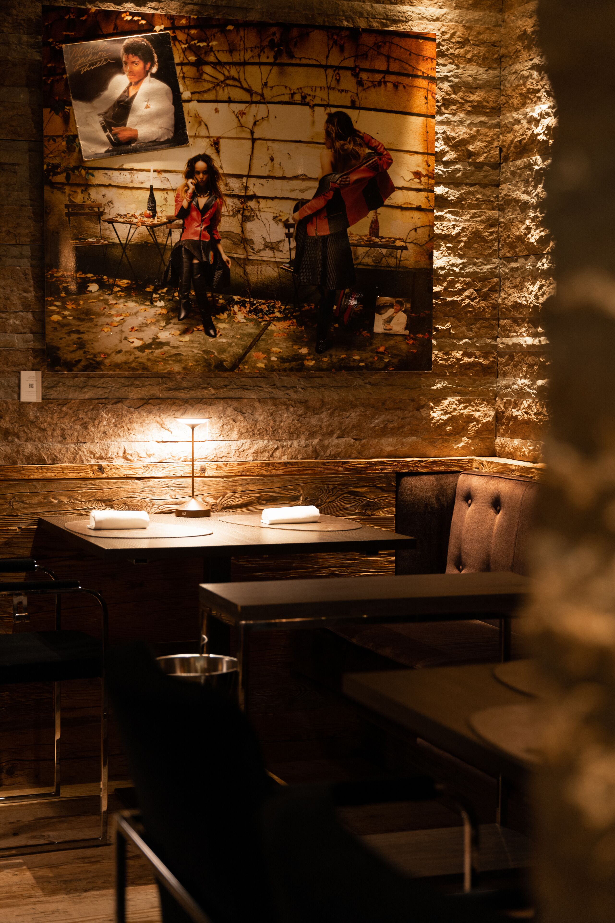 A dimly lit restaurant with a rustic stone wall, a table with a lamp, and a poster of a person on the wall.
