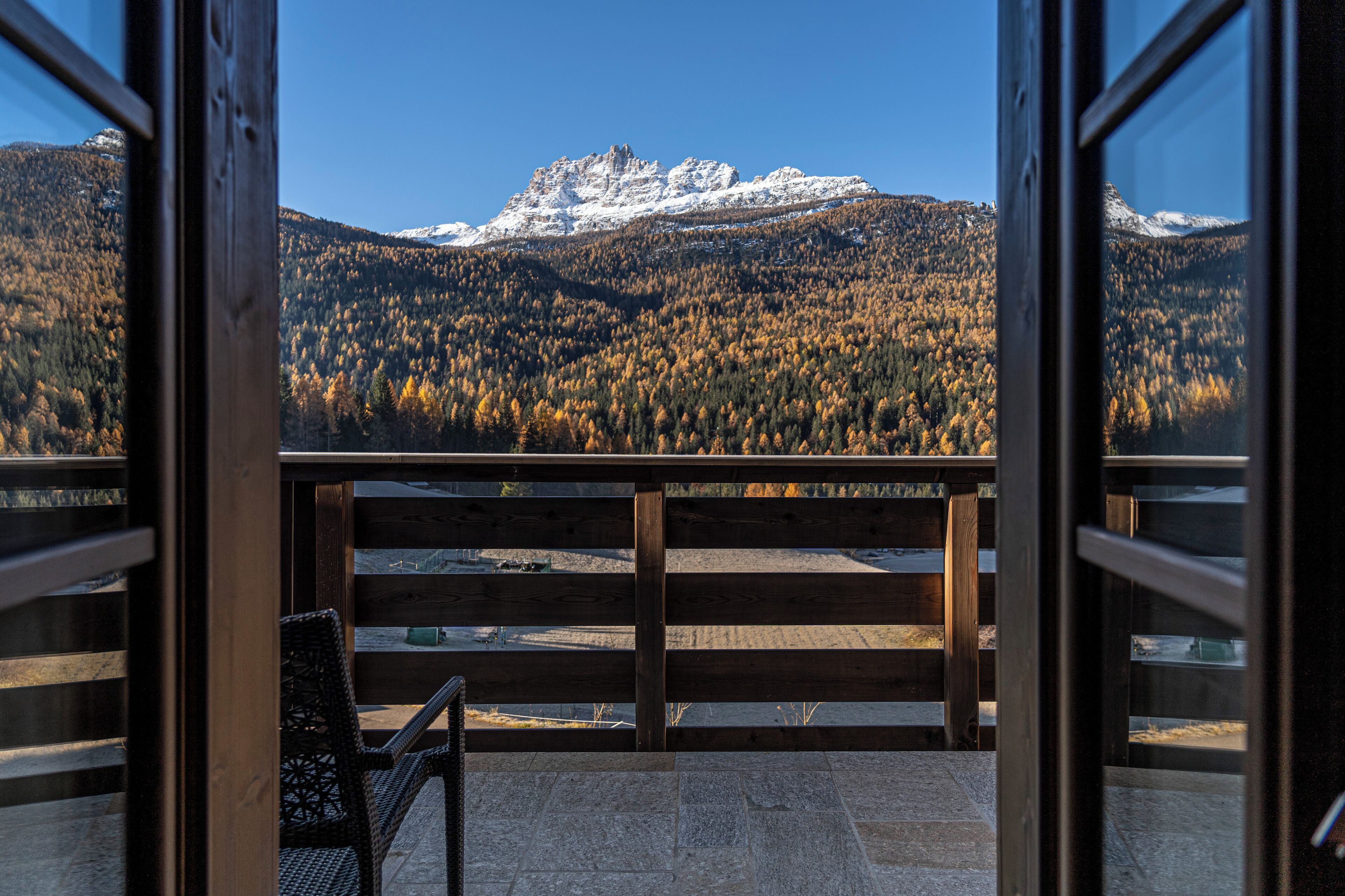 A balcony with a wooden railing overlooking a mountainous landscape with snow-capped peaks and autumn-colored trees.