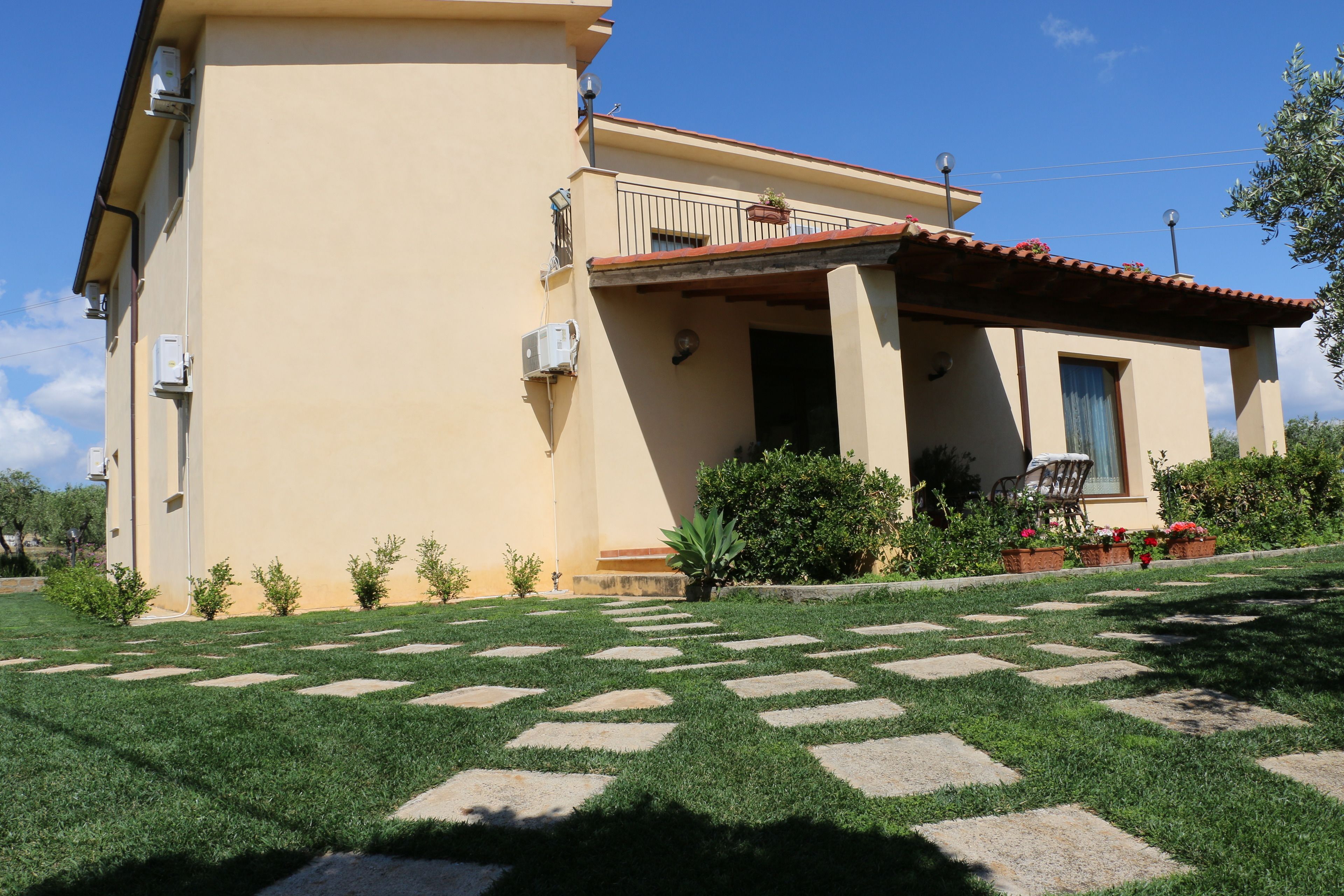 A two-story house with a balcony, a small garden with stepping stones, and a clear blue sky.