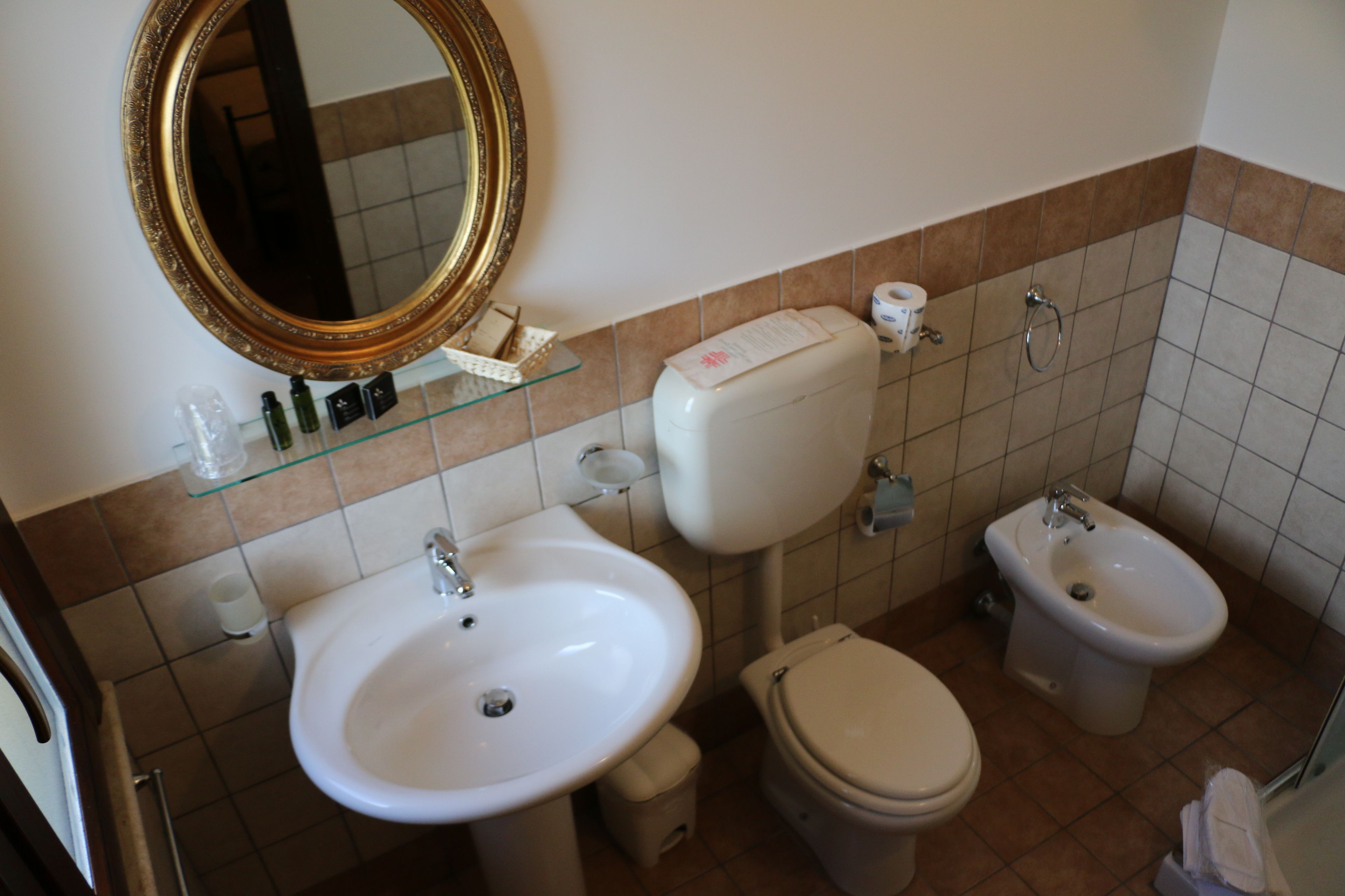 A bathroom with a white sink, toilet, bidet, and a large mirror with a gold frame.