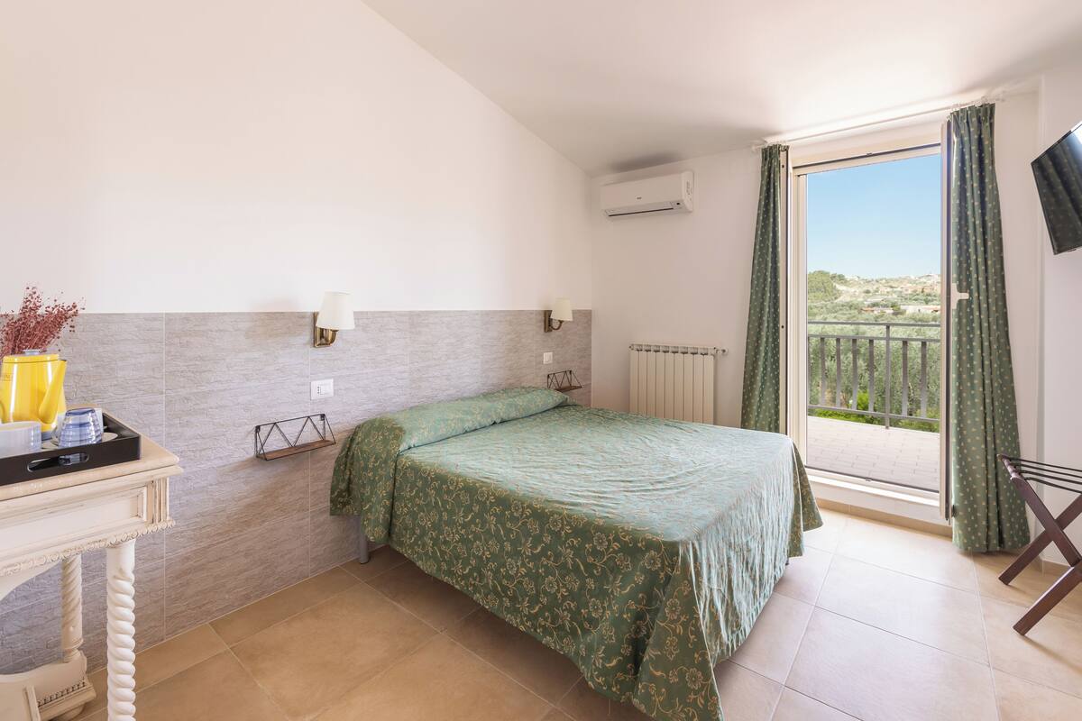 A bedroom with a bed, a nightstand, a lamp, a window with curtains, and a view of the outside.