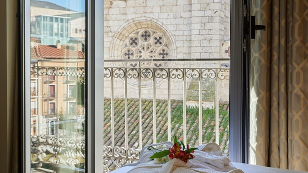 Une chambre d’hôtel avec un lit, une fenêtre donnant sur un immeuble orné d’une fenêtre décorative et un balcon avec une balustrade.