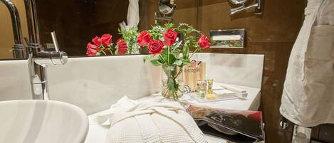 A bathroom with a white sink, a mirror, a towel, a vase with red roses, and toiletries on the counter.