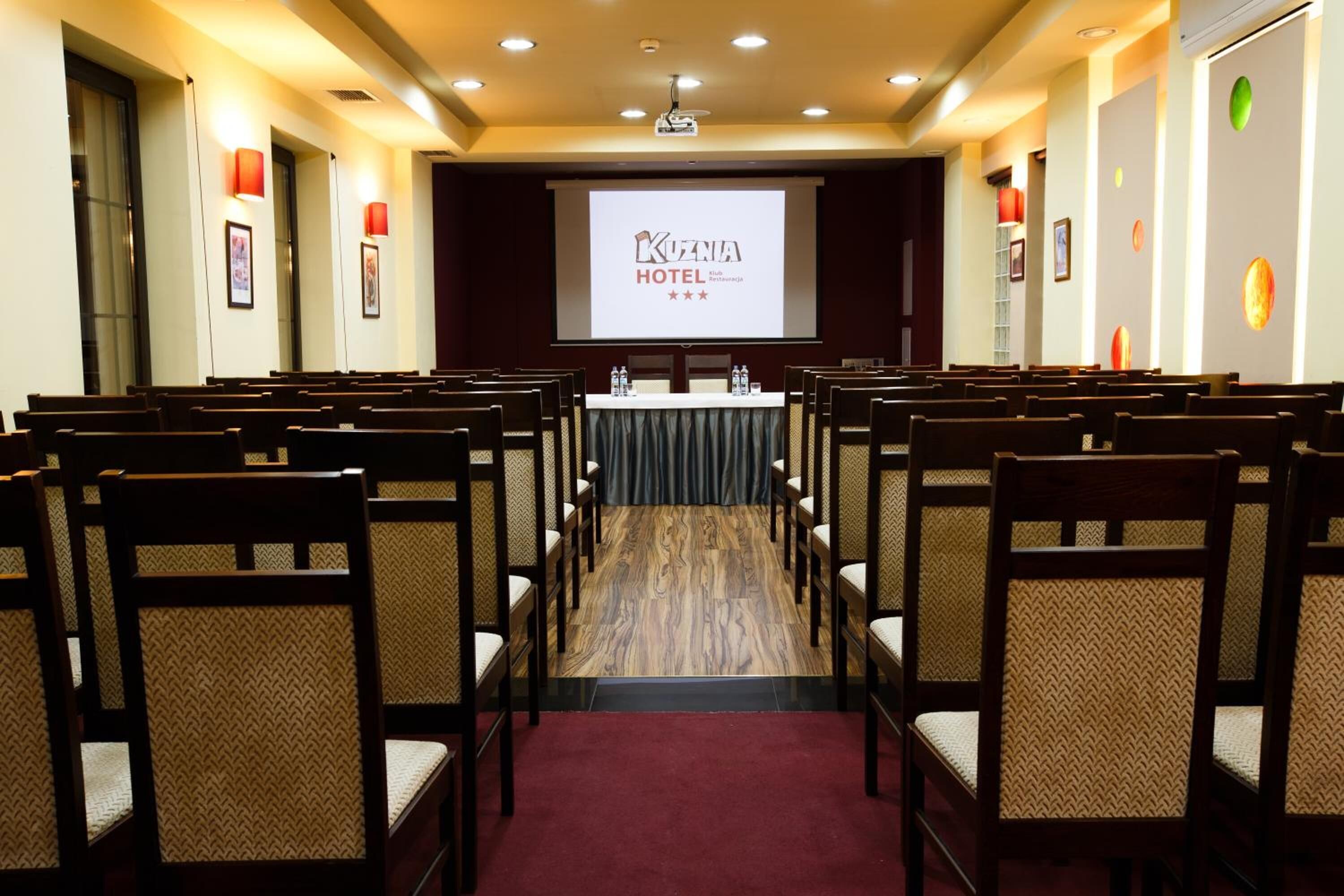 Ein Konferenzraum im Hotel Kuzenia mit Reihen aus Holzsesseln, einer Projektionsleinwand und einer Bühne.
