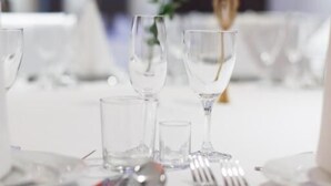 Une table à manger dressée avec de la vaisselle en verre transparent et des couverts, un vase contenant une seule fleur blanche et un objet décoratif doré.