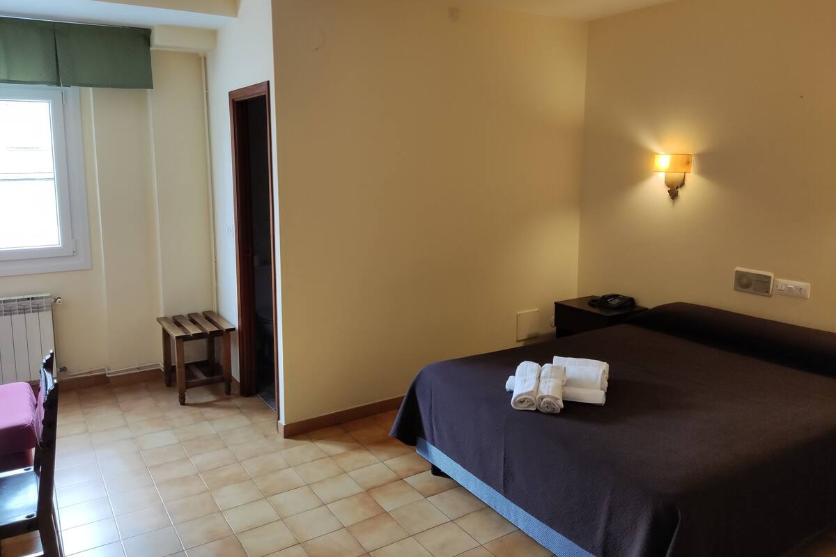 A hotel room with a bed, a desk, a chair, a radiator, and a wall-mounted light fixture.