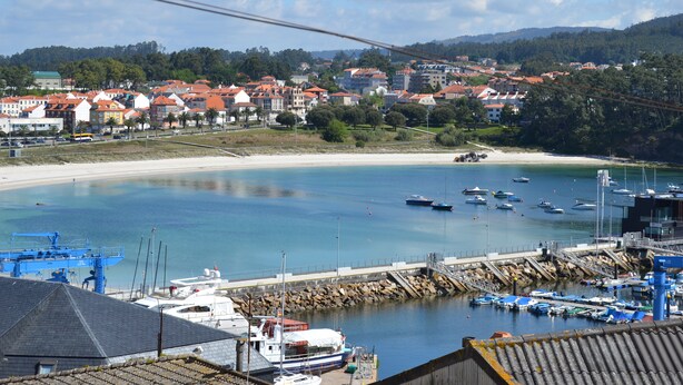 Un puerto deportivo costero con barcos amarrados, una playa de arena y edificios residenciales al fondo.
