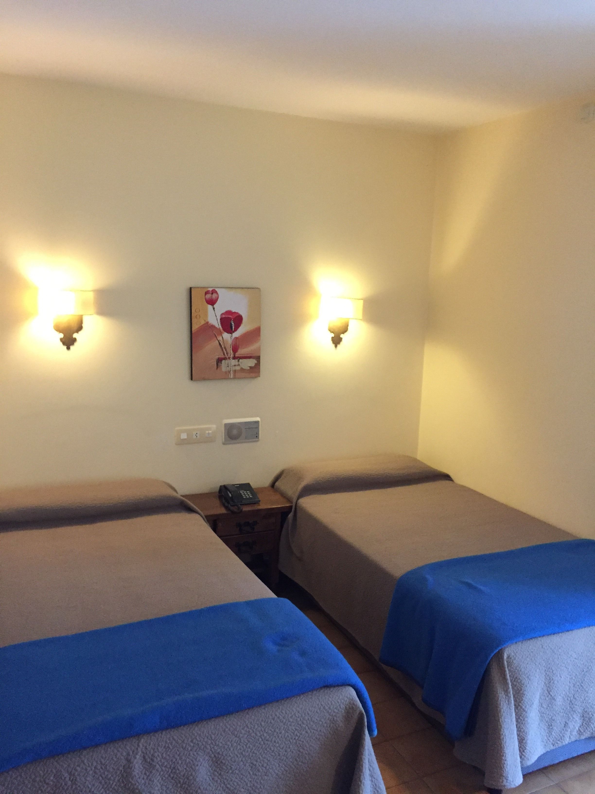 A hotel room with two beds, each with a blue blanket, a nightstand with a phone, and wall-mounted lamps.