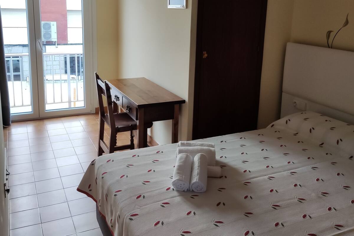 A hotel room with a bed, a desk, and a chair. There is a door and a window with a view of the outside.