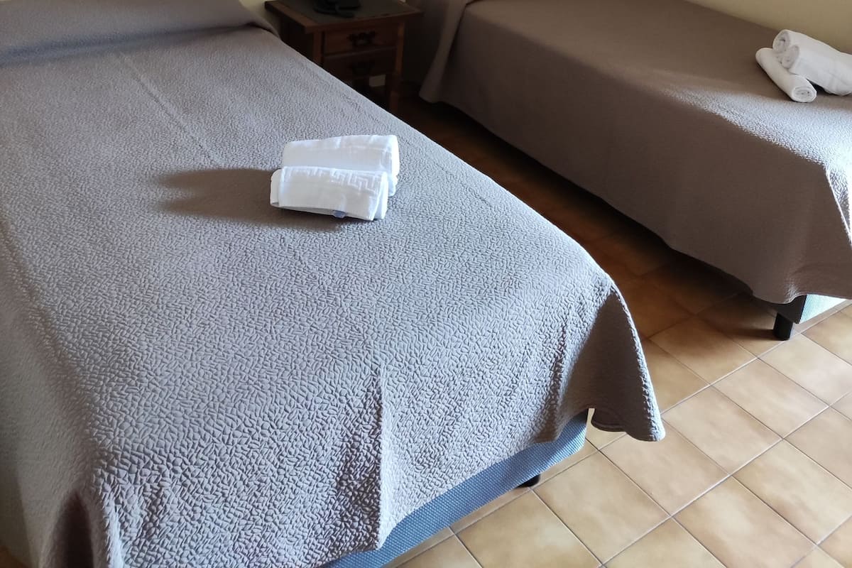 A hotel room with two single beds, a nightstand with a phone and a lamp, and a wall-mounted light fixture.
