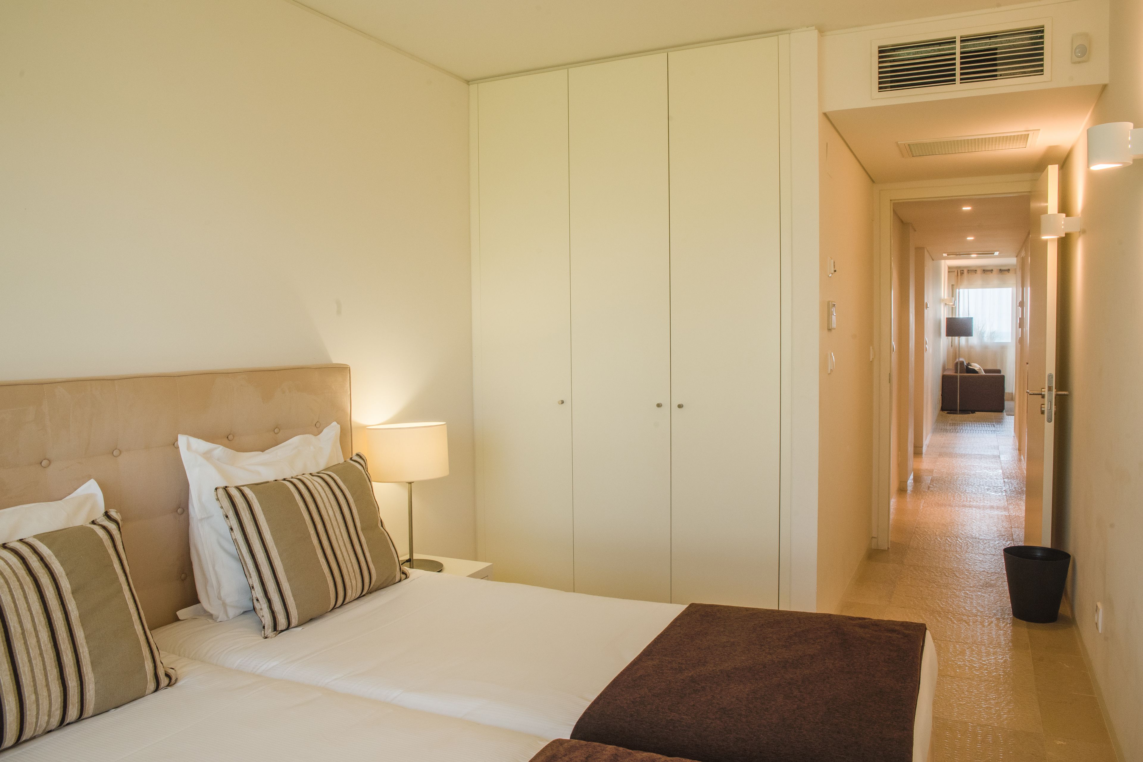 Habitación de hotel con cama grande, armario, mesita de noche con lámpara y vista al pasillo.