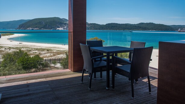 A wooden deck with a table and chairs overlooking a beach and turquoise sea. - Troia Residence Praia (Grandola)