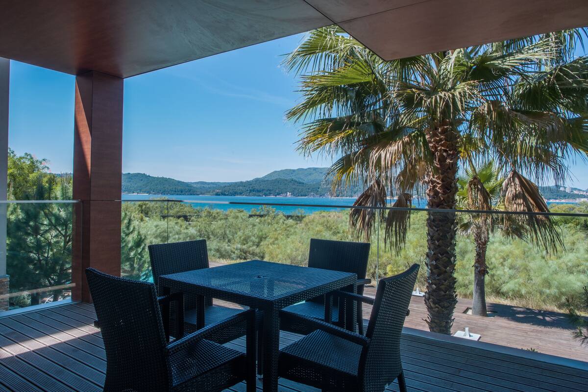 A terrace with a table and chairs, overlooking a body of water and hills.