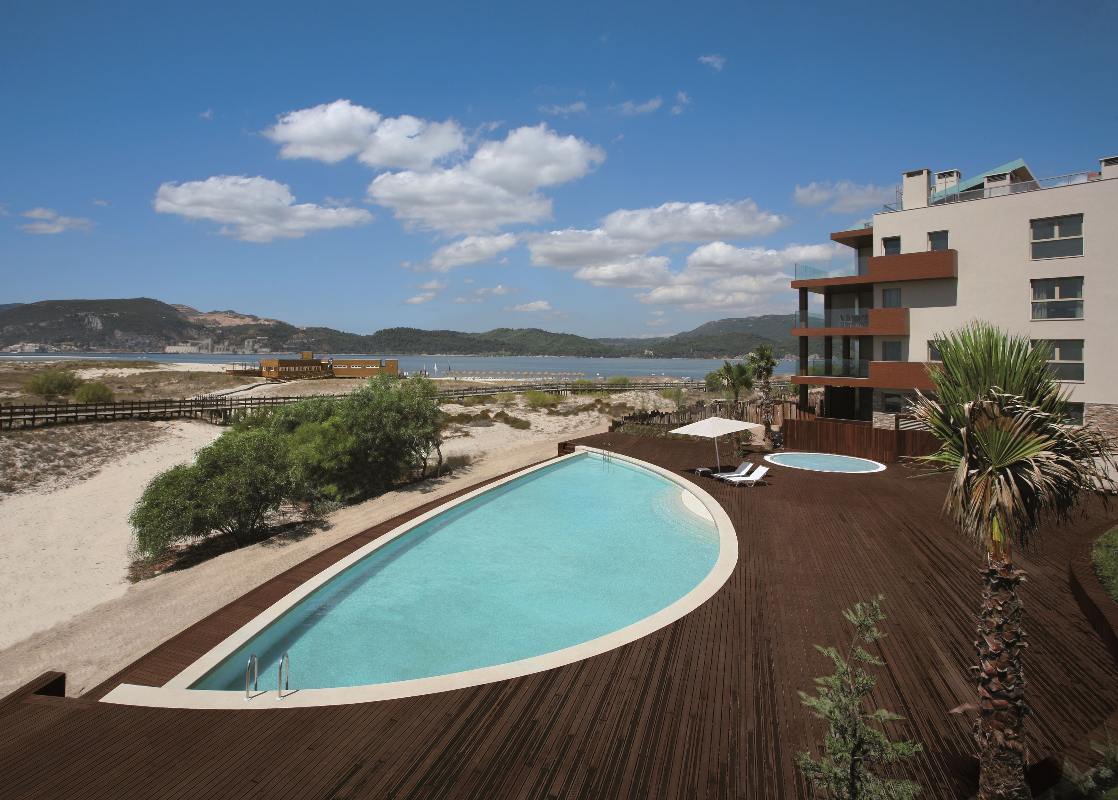 A pool area with a wooden deck, lounge chairs, and a view of the beach and sea.