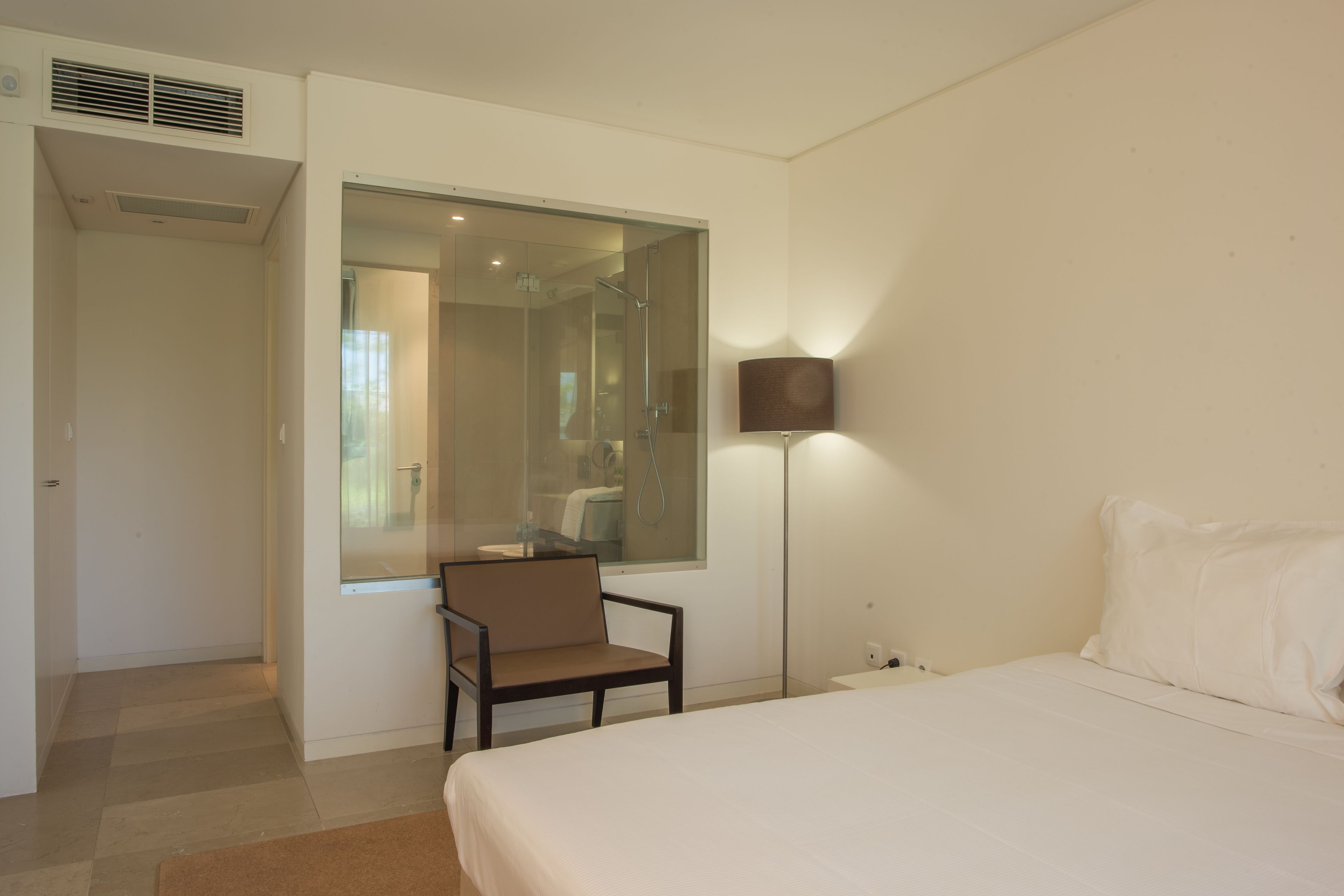 Une chambre à coucher comprenant un lit, une chaise, un grand miroir, une lampe sur pied et une salle de bain visible à travers la porte en verre.