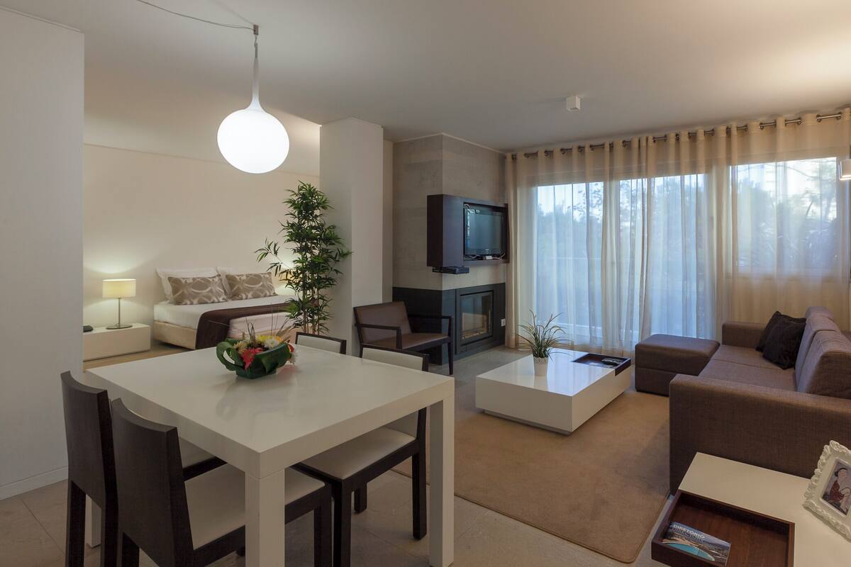 A modern living room with a dining area, a flat-screen TV mounted above a fireplace, and a large window with sheer curtains.
