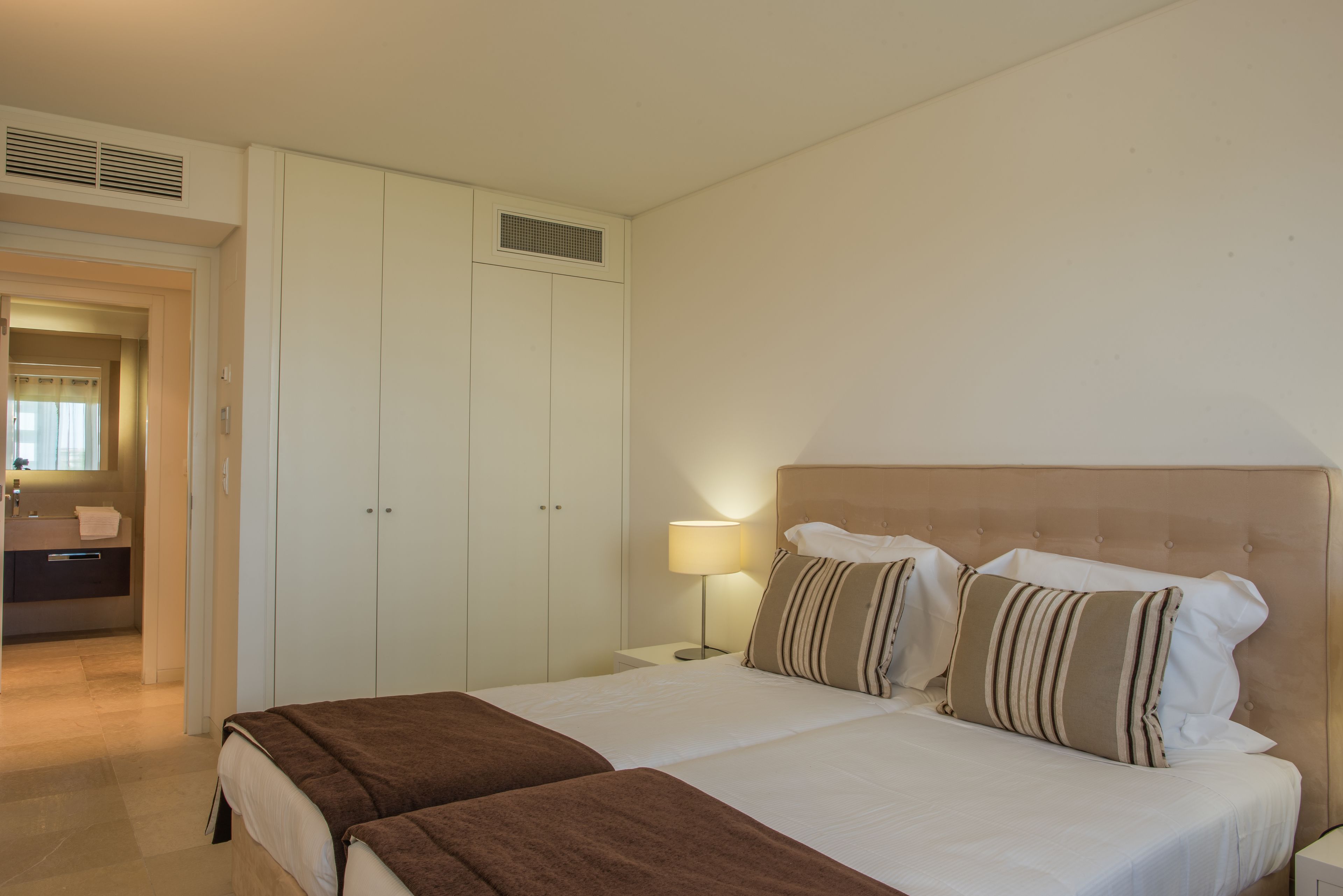 Quarto de hotel com uma cama grande, um guarda-roupa, uma mesa de cabeceira com luminária e um banheiro visível através de uma porta aberta.